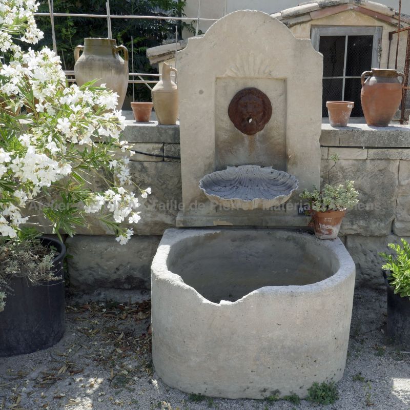 Fontaine murale en pierre avec une sortie d'eau représentant une tête de lion qui contraste avec la couleur blanchâtre de la pierre naturelle.
